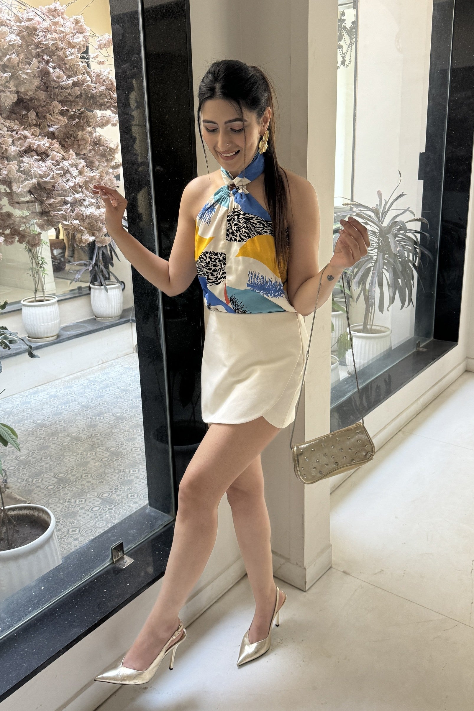 A woman standing indoors wearing a sleeveless top with a vibrant multicolor geometric print, consisting of yellow, blue, and gray tones, paired with a beige skirt.