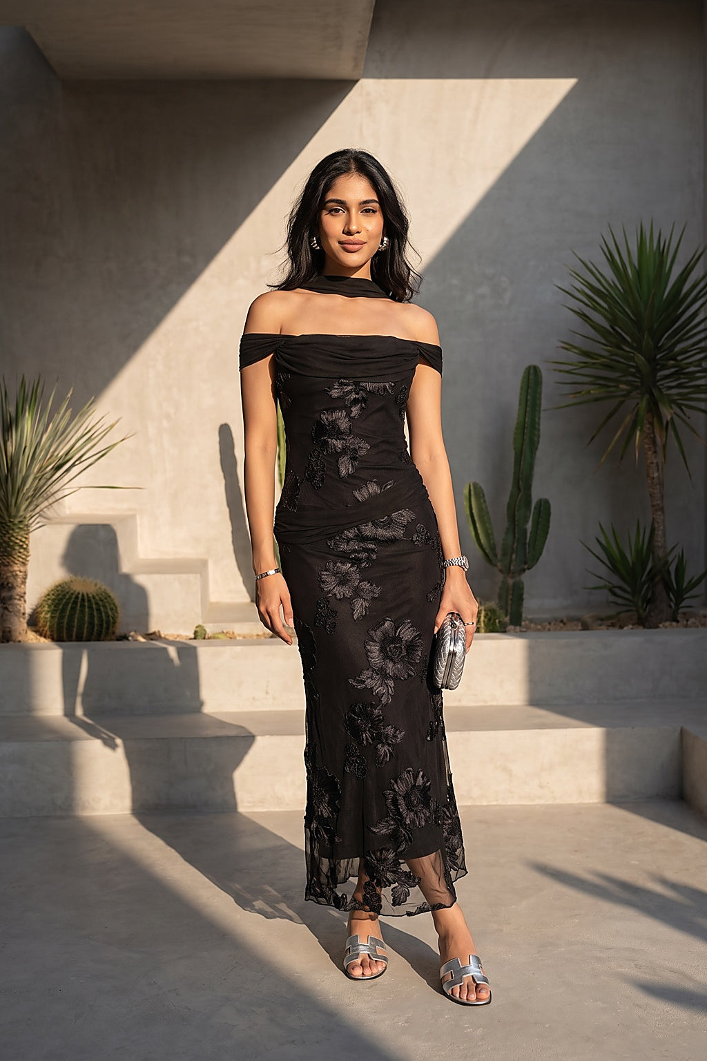 A woman stands outdoors in front of a modern concrete wall and steps, wearing a black off-the-shoulder lace maxi dress with a sheer floral pattern. She is holding a small metallic clutch and wearing silver heeled sandals.
